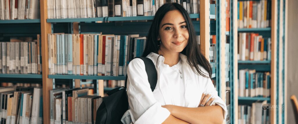 College-aged girl standing in the library in front of a bookshelf.