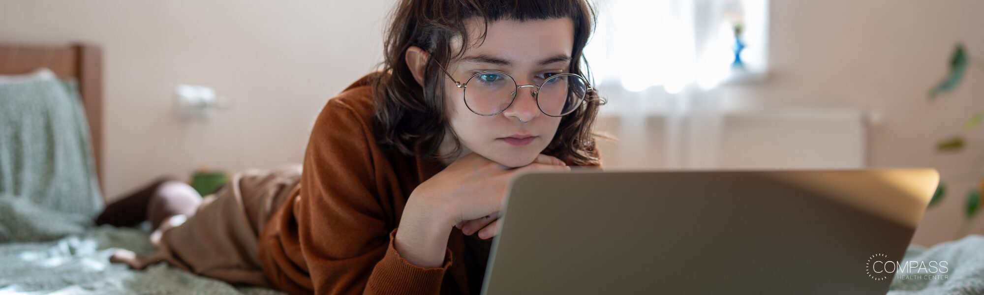 A young woman with curly dark hair and round glasses lies on a bed, resting her chin on her hand while looking intently at a laptop screen. She wears a rust-colored sweater, and soft natural light comes through a window in the blurred background.