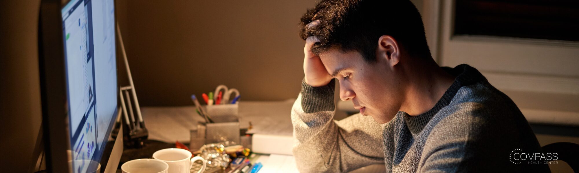 A young man sits at a desk late at night, head resting in his hand, looking down with a stressed expression. His computer screen glows beside him, and the desk is scattered with mugs, pens, and papers. Compass Health Center logo in the bottom corner.