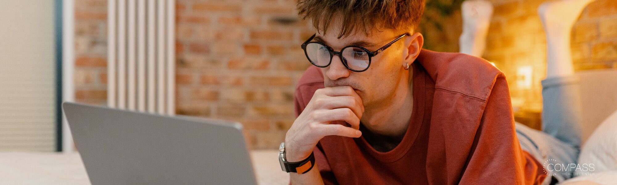 A young person with glasses and a rust-colored sweater lies on a bed, resting his chin thoughtfully on his hand as he looks at a laptop screen. He's in a room with an exposed brick wall and warm lighting in the background.