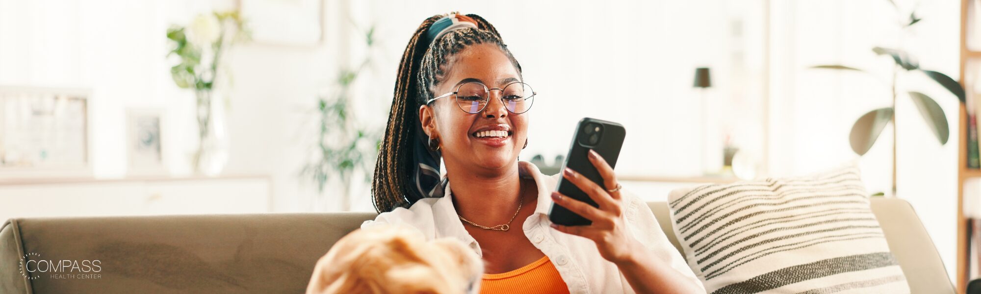 A young woman with braided hair and glasses smiles while looking at her phone, sitting on a couch in a bright, plant-filled living room with a striped throw pillow beside her and a dog partially visible in front of her.