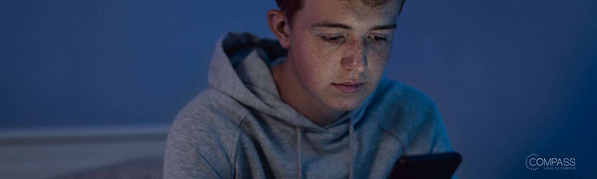 A young man in a gray hoodie looks down at his phone in a dimly lit room, his face lit by the phone's glow against a blue-toned background.