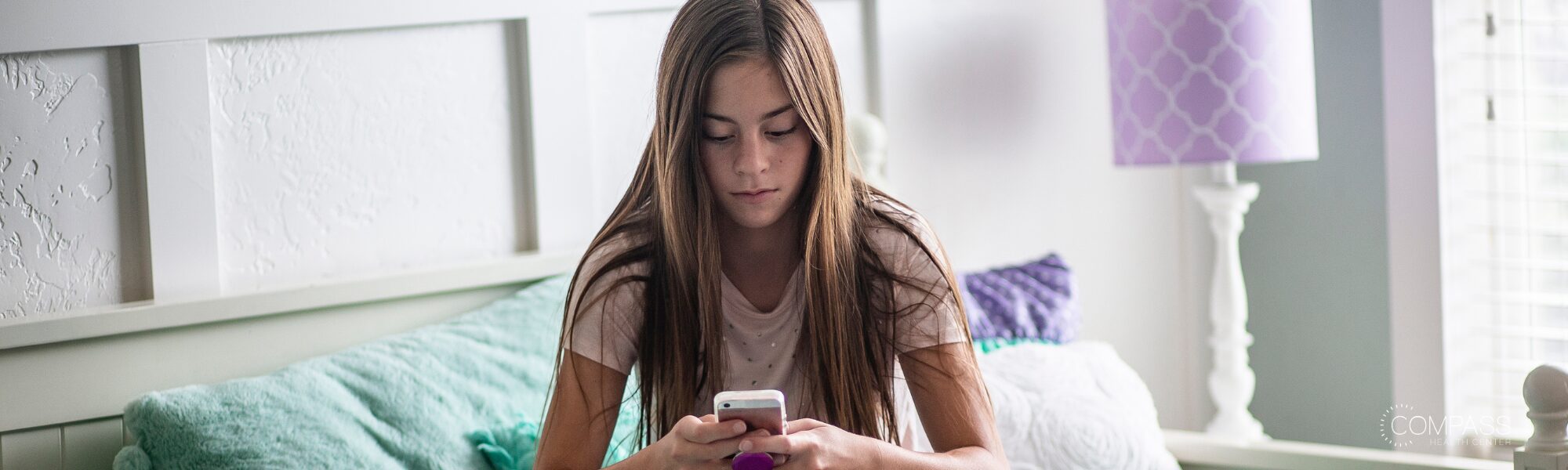 A young girl with long brown hair sits on a bed with teal and purple pillows, looking down at her phone with a serious expression. A white headboard and a purple lamp are visible in the softly lit bedroom behind her.