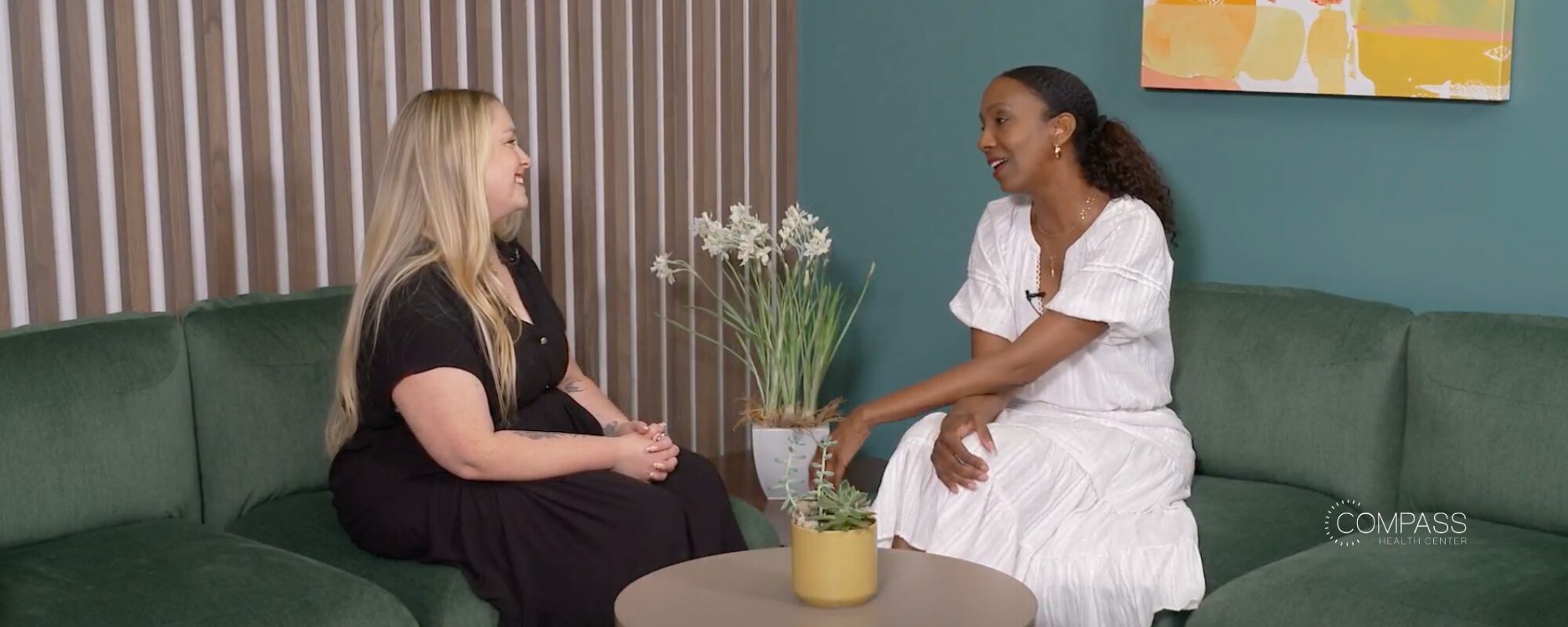 Two women sit facing each other in conversation on a green sofa, in a warm, softly lit room with a striped wood accent wall, a teal wall with an abstract painting, and a small round table between them holding potted plants. One woman, with long blonde hair, wears a black dress; the other, with curly dark hair, wears a white dress and gestures as she speaks. The Compass Health Center logo appears in the bottom right corner.