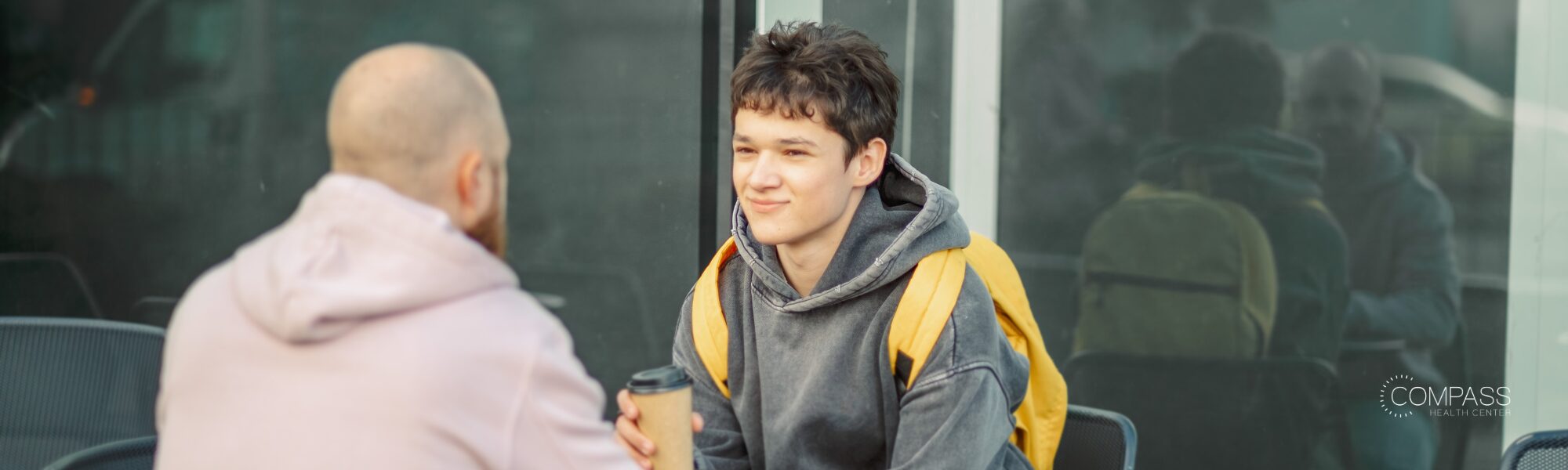 A teenage boy wearing a gray hoodie and yellow backpack sits outdoors talking with an adult holding a coffee cup, with a glass building reflecting the scene behind them. The Compass Health Center logo appears in the bottom right corner.