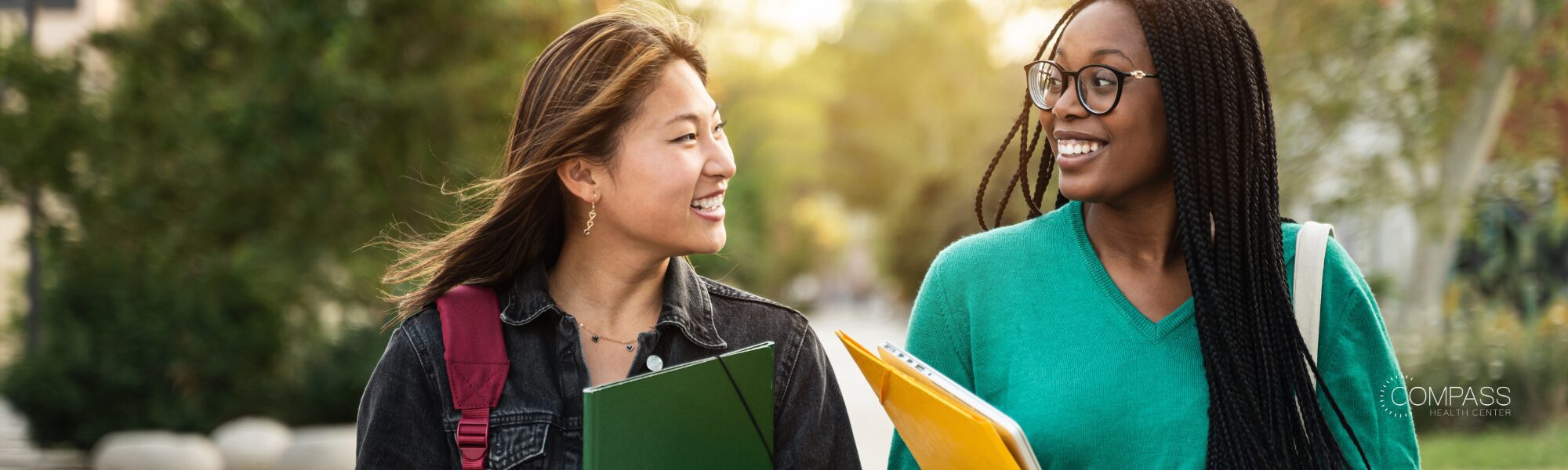 Two young women walk together outdoors on a tree-lined path, smiling and talking, one carrying a backpack and folder, the other wearing glasses and carrying a folder. The Compass Health Center logo appears in the bottom right corner.