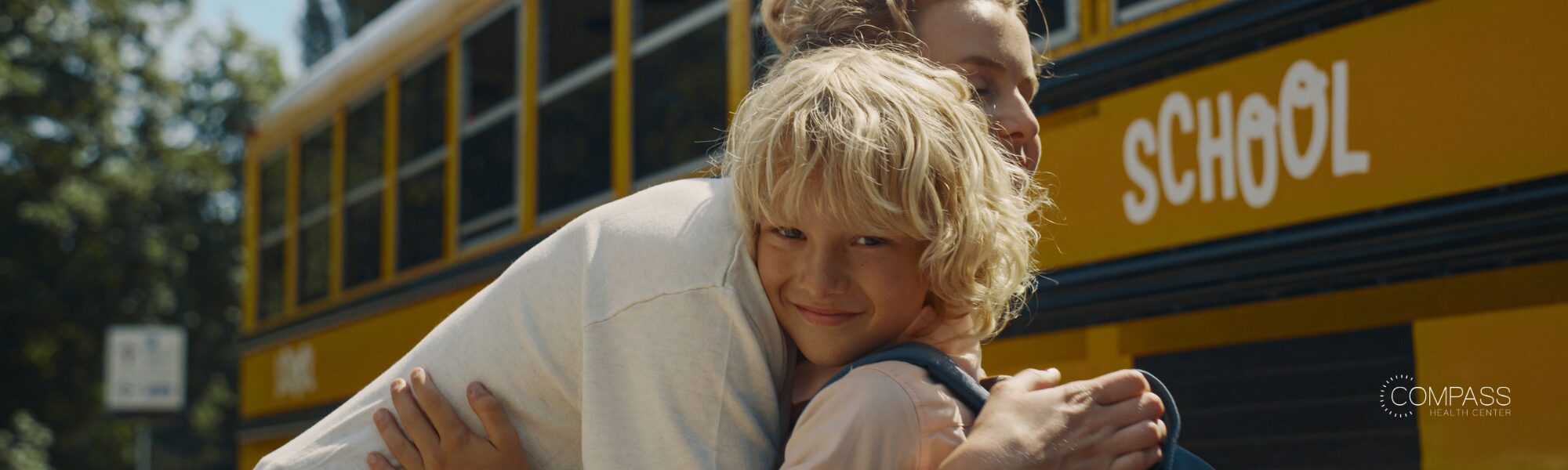 A young boy with curly blond hair smiles while hugging an adult tightly in front of a yellow school bus. The Compass Health Center logo appears in the bottom right corner.