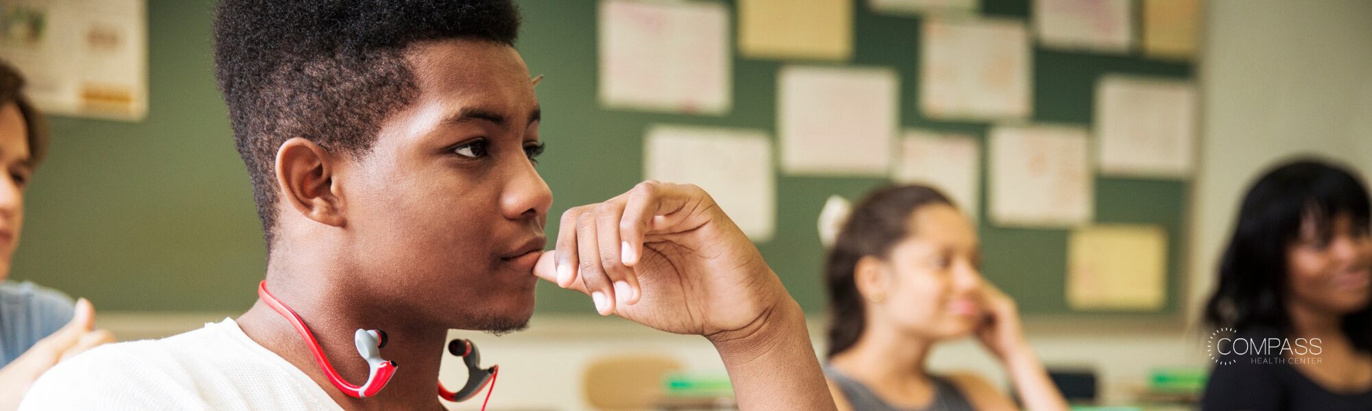 A teenage boy with neck-style wireless earphones sits thoughtfully in a classroom, resting his fingers near his mouth, with classmates visible blurred in the background near a bulletin board. The Compass Health Center logo appears in the bottom right corner.