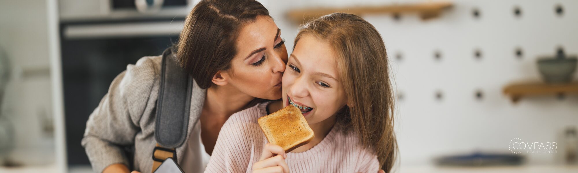 A mother wearing a backpack kisses her smiling teenage daughter, who has braces, on the cheek while eating a piece of toast in a kitchen. The Compass Health Center logo appears in the bottom right corner.