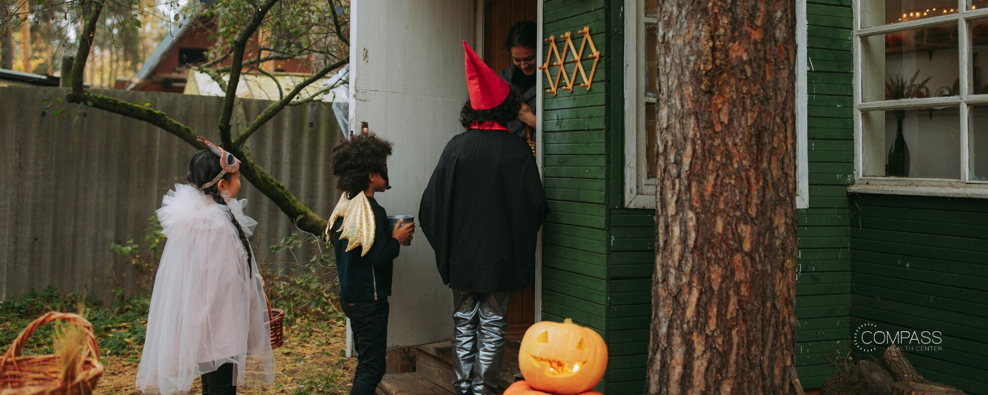 Three children in Halloween costumes trick-or-treating at the door of a green house, with a carved jack-o’-lantern on the porch step. Compass Health Center logo in the lower right.