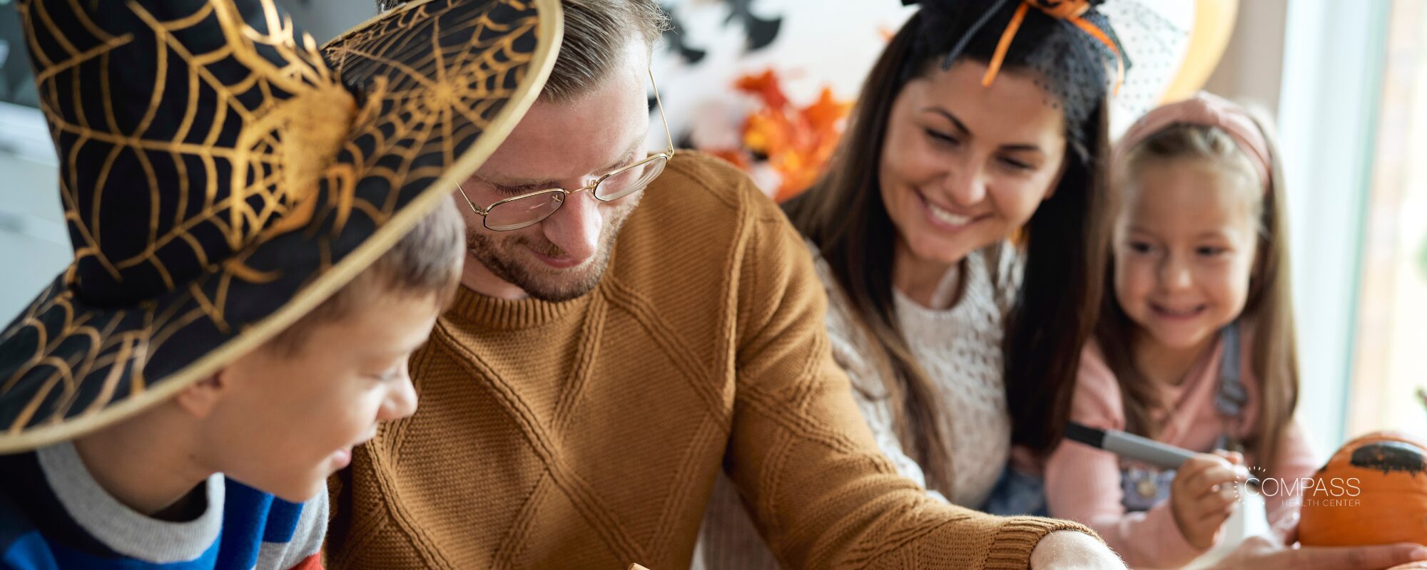 A family decorating pumpkins together. A boy in a spiderweb witch hat and a man in glasses look down at their project while a smiling woman and a young girl holding a marker look on. Compass Health Center logo in the lower right.