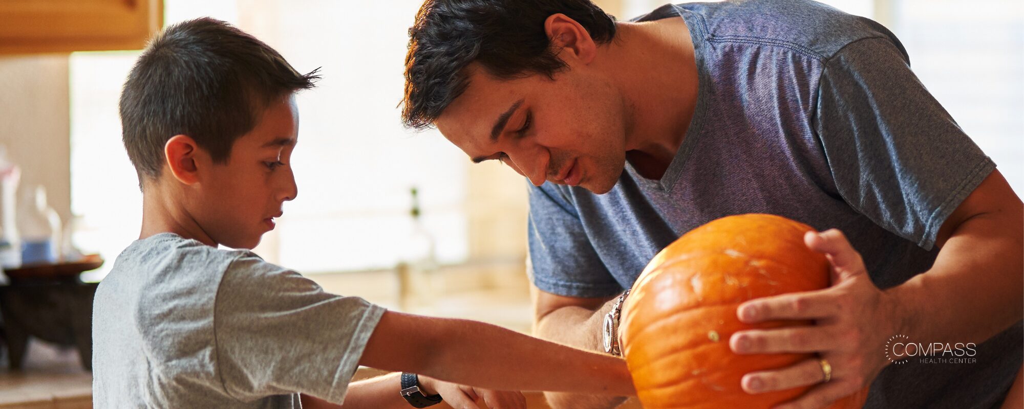A man and a boy leaning over a large orange pumpkin on a kitchen counter, getting ready to carve it. Compass Health Center logo in the lower right.