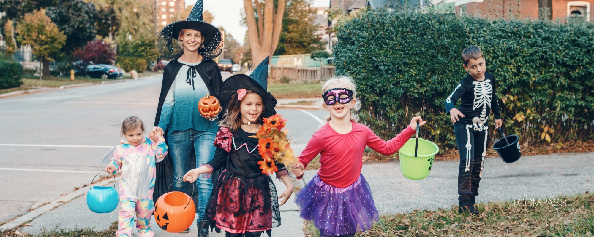 A woman in a witch costume walks down a residential street with four children in Halloween costumes, including a skeleton outfit, a purple tutu with a sparkly mask, and a witch hat, carrying plastic pumpkin buckets. Compass Health Center logo in the lower right.