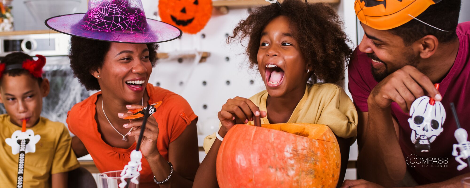 A laughing family of four decorating Halloween treats and a carved pumpkin at a kitchen table, wearing witch and pumpkin hats and holding skeleton-themed straws. Compass Health Center logo in the lower right.