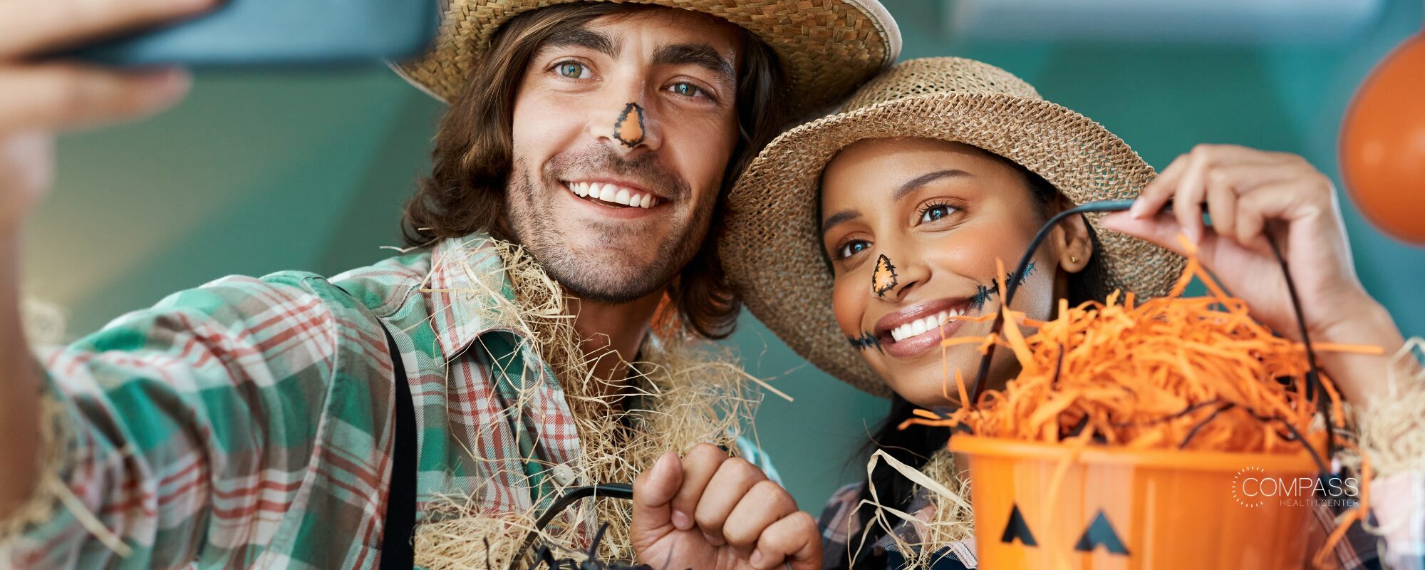 A smiling man and woman dressed as scarecrows in straw hats with painted noses, taking a selfie. She holds an orange jack-o’-lantern bucket filled with orange paper shreds. Compass Health Center logo in the lower right.