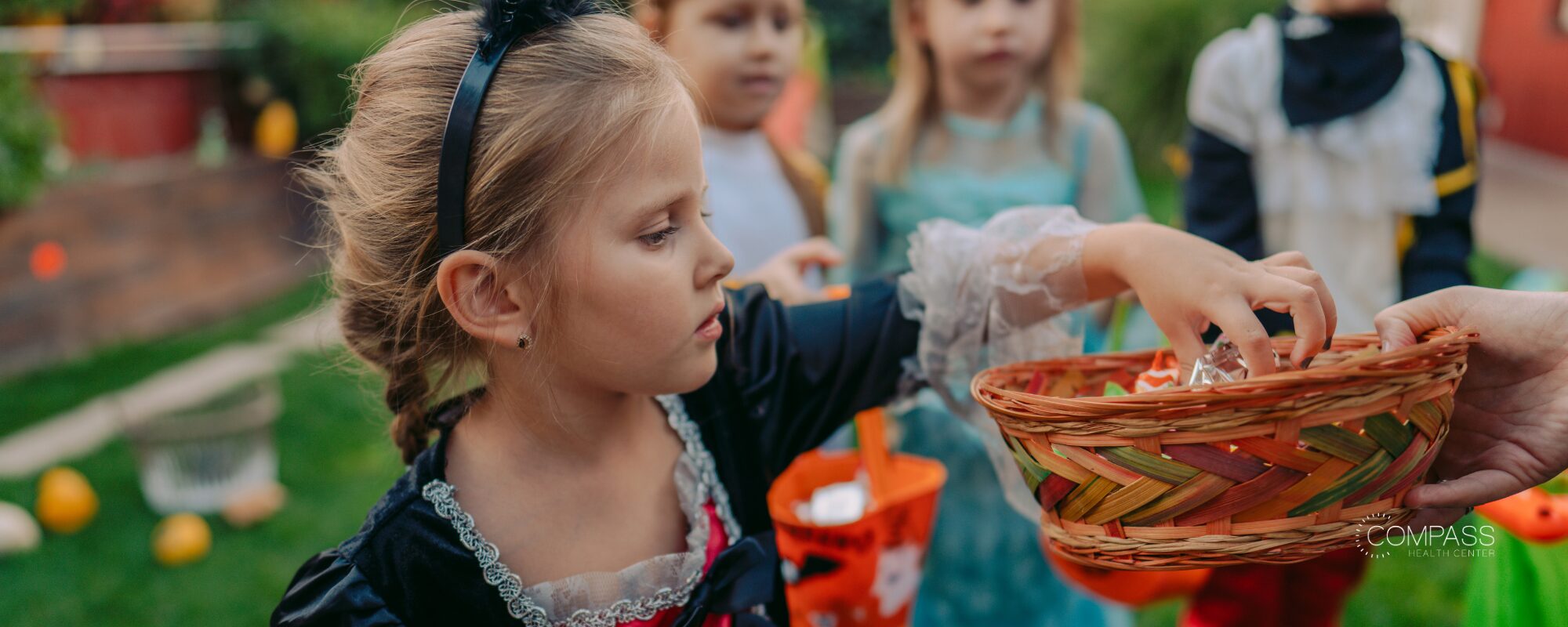 A young girl in a costume reaches into a wicker basket of candy held out by an adult, with other costumed children waiting behind her. Compass Health Center logo in the lower right.