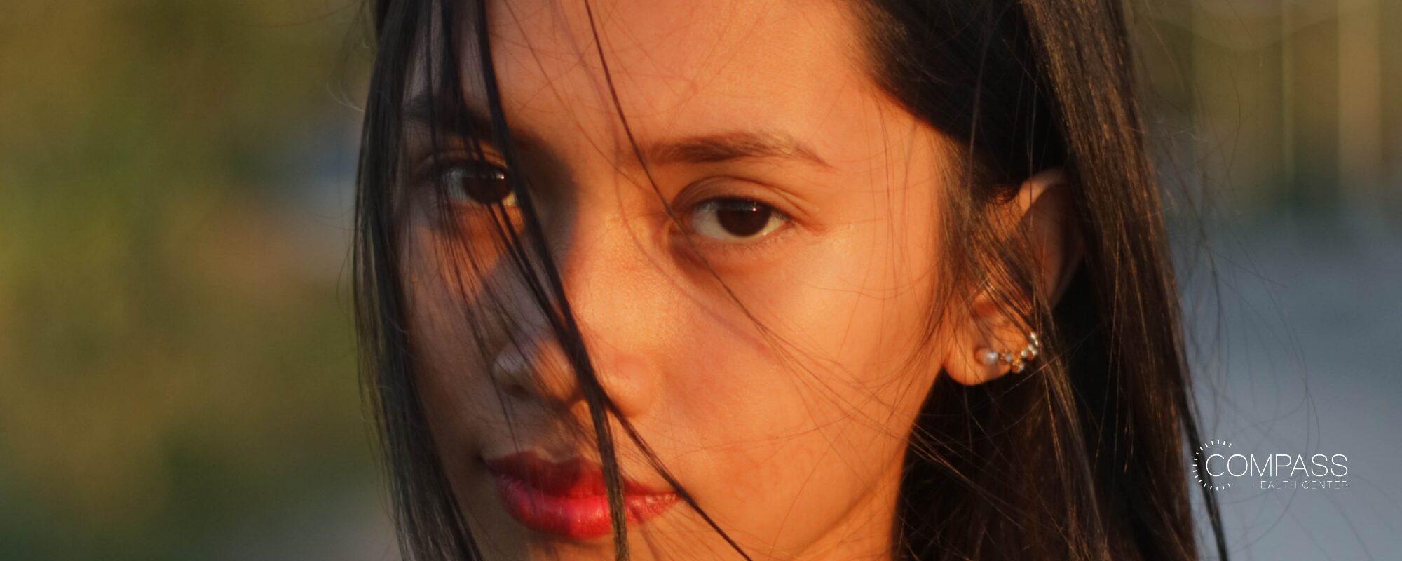 Close-up of a young woman with long dark hair partly covering her face, looking directly at the camera in warm golden light. Compass Health Center logo in the lower right.