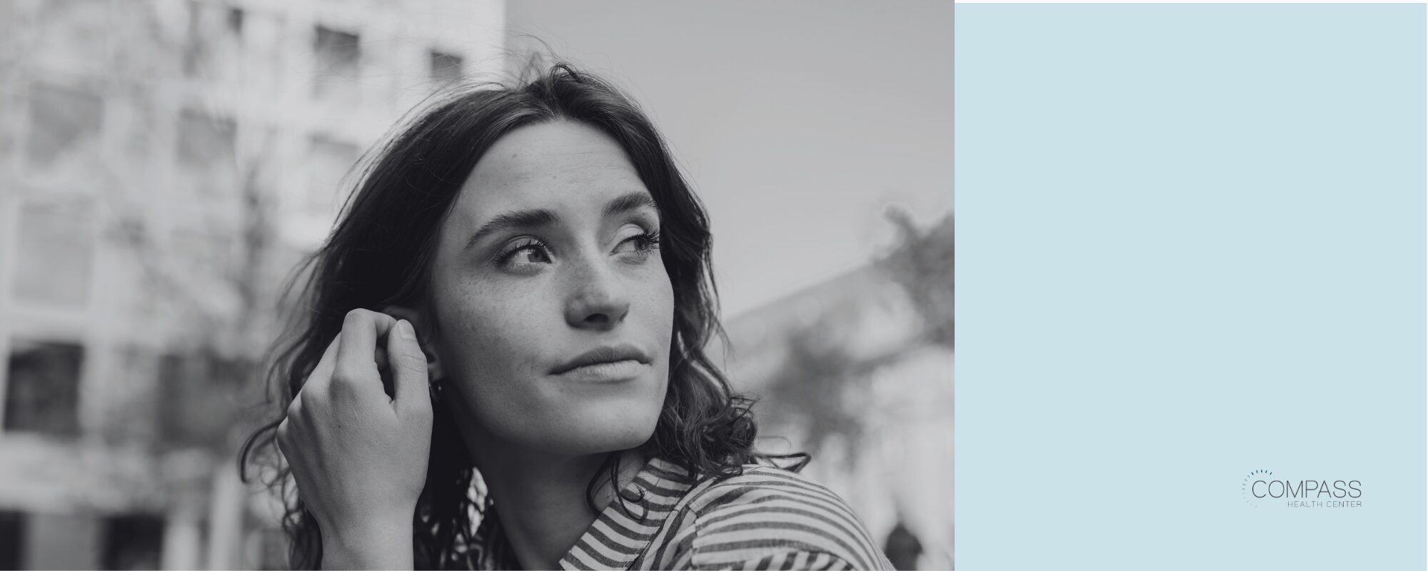 A black-and-white photo of a young woman with wavy dark hair and a striped collared shirt, gazing thoughtfully off to the side with one hand raised to her ear, in front of a blurred city building. A solid light blue panel fills the right side of the image, with the Compass Health Center logo in the bottom right corner.
