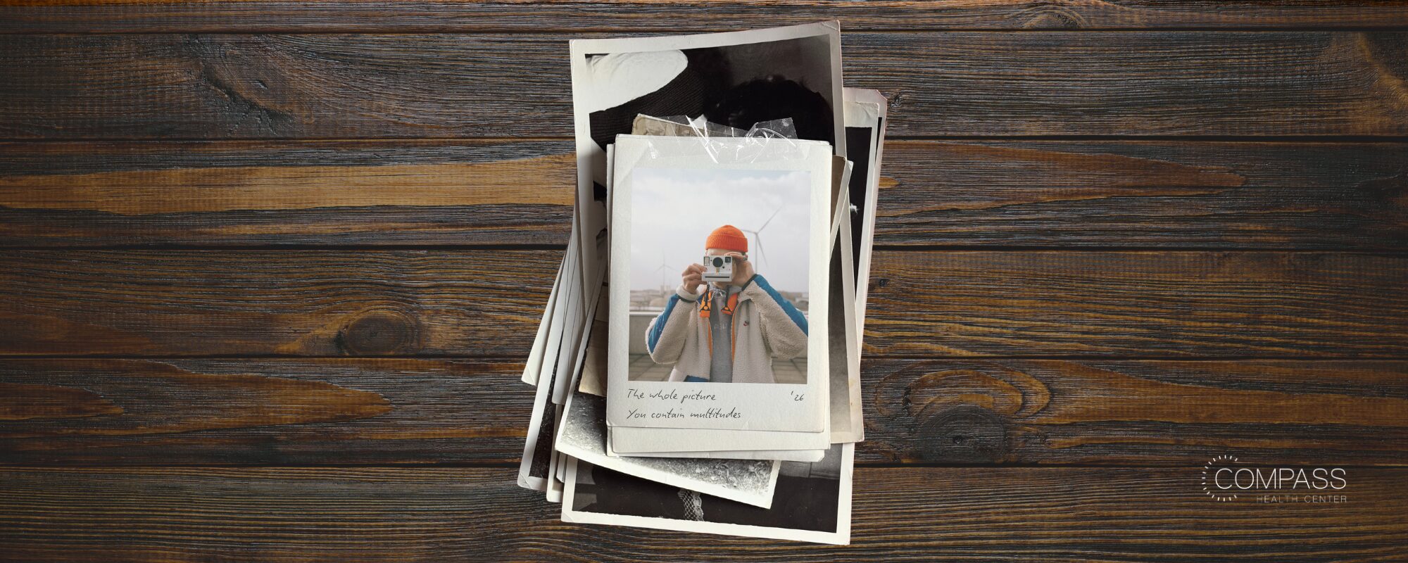 A stack of overlapping Polaroid-style photos on a dark wood surface. The top photo shows a person in an orange beanie and fleece jacket holding a camera up to their face, with wind turbines in the background. Handwritten captions read "The whole picture," "You contain multitudes," and "'26." The Compass Health Center logo appears in the bottom right